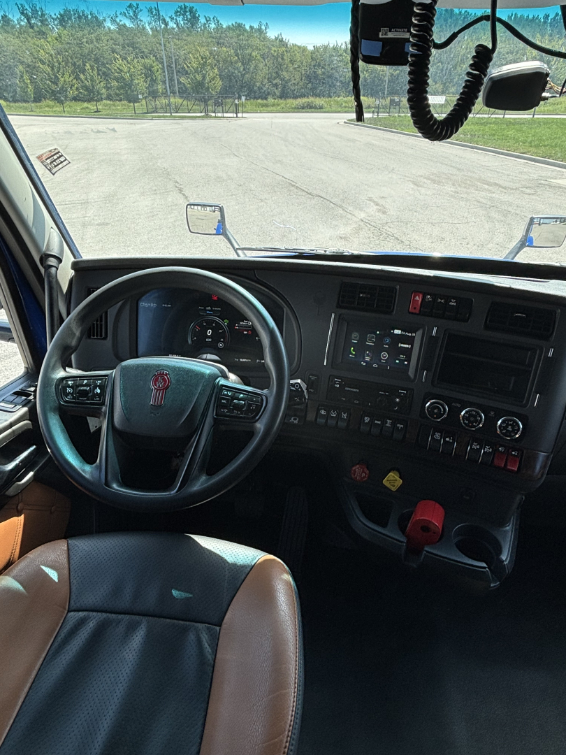Cab interior of a 2023 Kenworth T680 showing steering wheel, dashboard with control panel and gear shifter, front windshield and side mirrors.