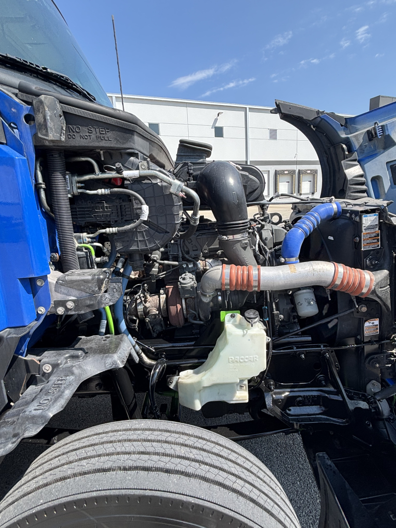 Truck engine bay of a 2023 Kenworth T680 showing air intake, coolant reservoir, hoses, and engine components visible between the front wheels.