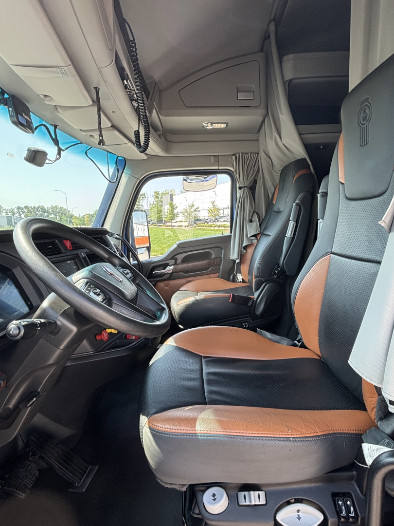 Kenworth T680 truck interior showing driver seat and passenger seat with tan-brown and black vinyl; dashboard and steering wheel visible; overhead cabinet; windshield with side mirror; no visible labels or text.