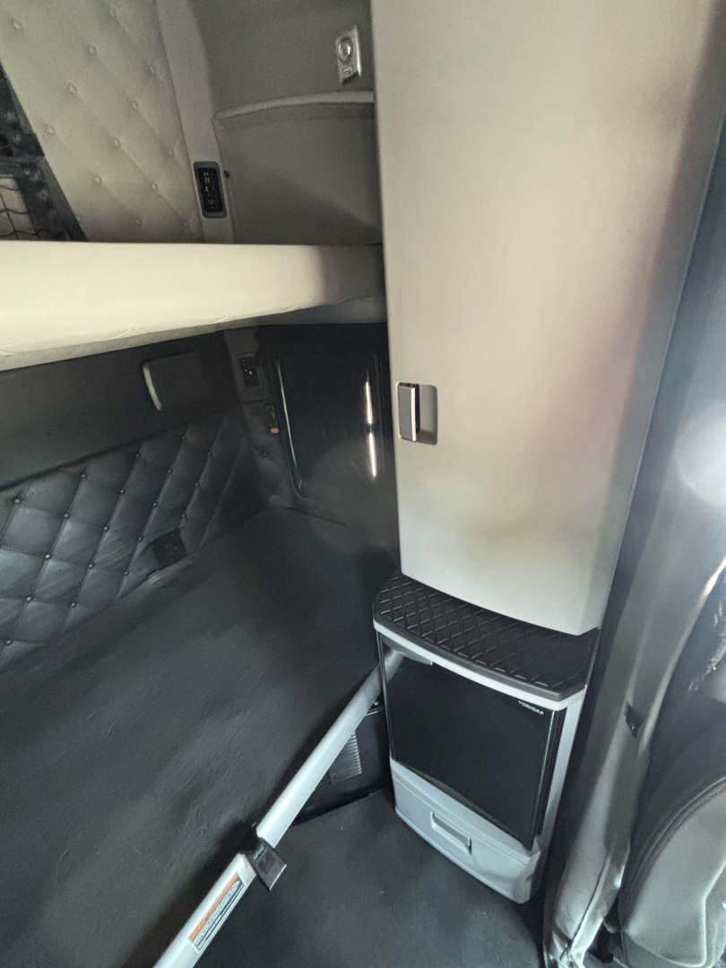 Kenworth T680 cab interior showing sleeper area and storage cabinet; vertical console with small step and panel, dark bedding, and metallic surfaces.