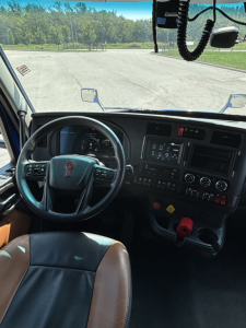 Cab interior of a 2023 Kenworth T680 showing steering wheel, dashboard with control panel and gear shifter, front windshield and side mirrors.