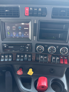 Kenworth T680 interior dashboard showing steering wheel area, climate controls, radio/navigation display, and various labeled switches.