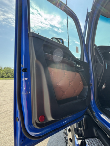 Door panel of a 2023 Kenworth T680 truck showing blue exterior, brown diamond-stitched door interior panel, open door, sky visible.