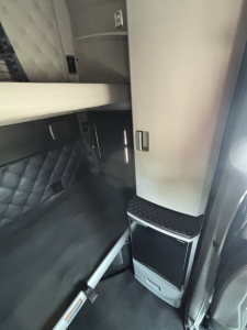 Kenworth T680 cab interior showing sleeper area and storage cabinet; vertical console with small step and panel, dark bedding, and metallic surfaces.