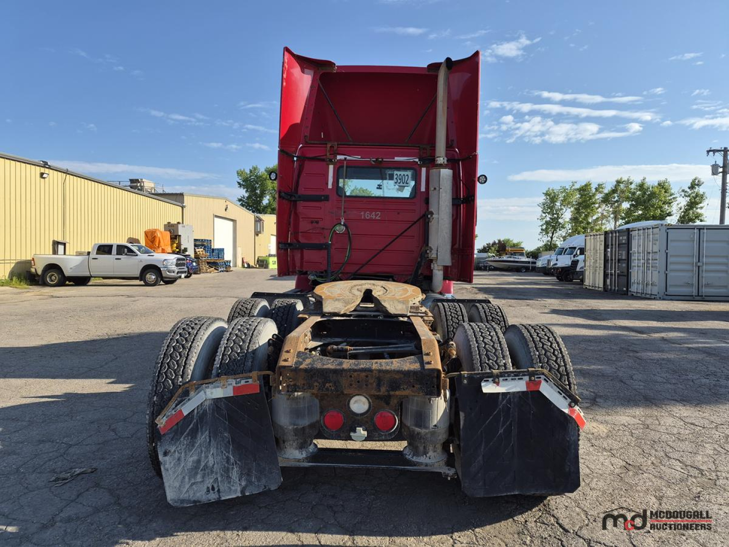 Volvo day cab semi truck rear view showing fifth wheel, dual rear axles, mud flaps, and red cab in background.