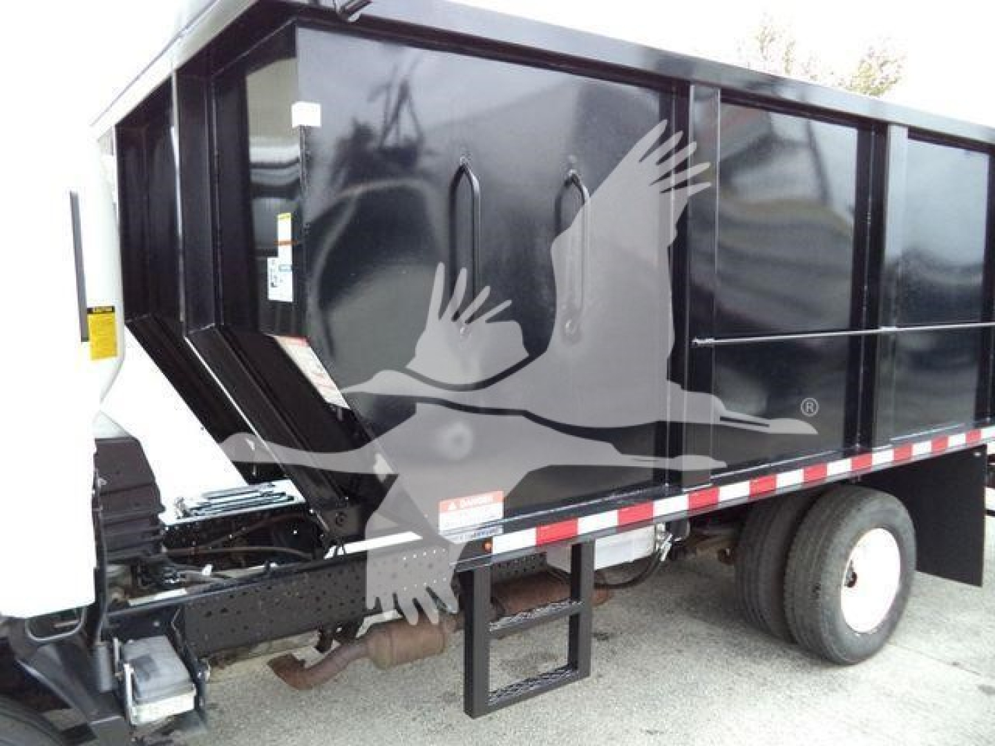 Isuzu NPR dump truck side showing truck bed and rear wheel; visible text includes none legible, but truck features a black dump body with white cab front and reflective tape along bed.