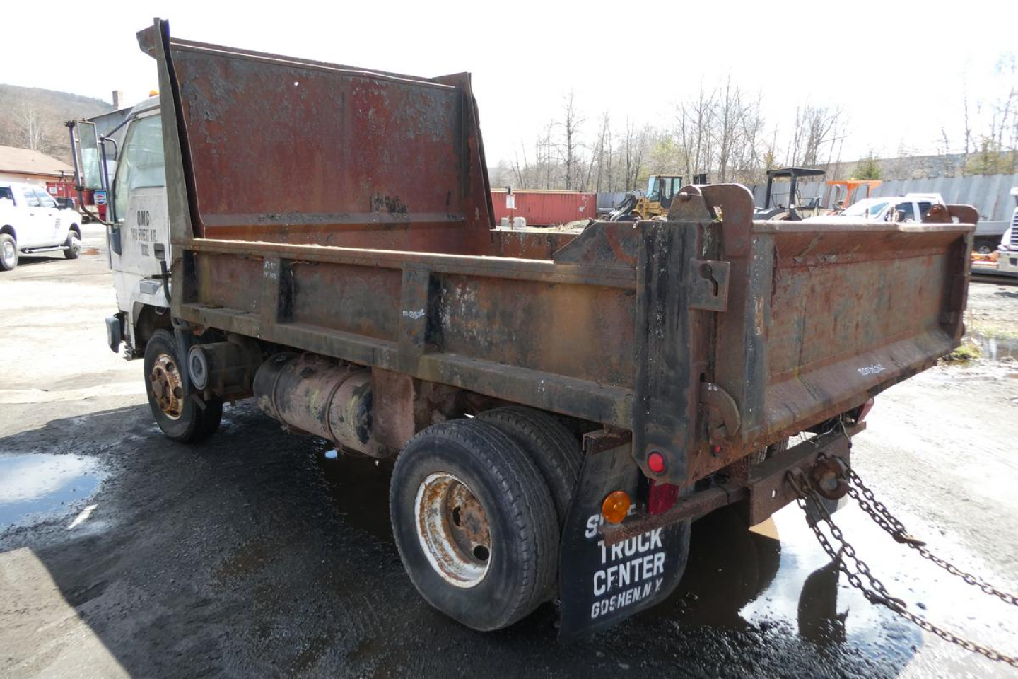 Dump truck bed showing rusted metal dump body and dual rear wheels with chain hitch and mud flaps.