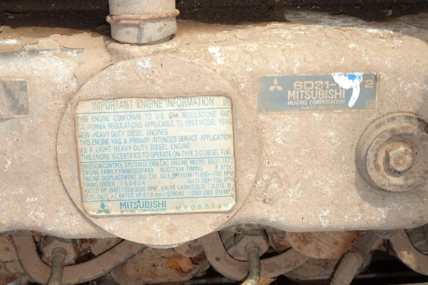 Mitsubishi diesel engine area with two blue labels. Left label has small text about engine information; right label shows 6031 MITSUBISHI MII... linkage and circular component with bolt. Visible text includes 'MITSUBISHI' and '6031' and 'MI