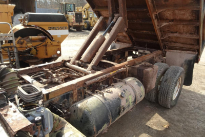 Dump truck chassis showing hydraulic pump and fuel tank with exposed frame and rear wheels under bed.