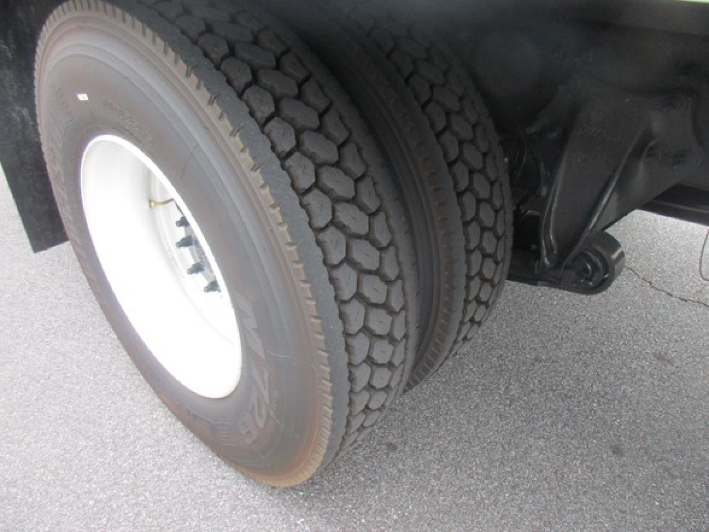 Truck rear wheel and suspension showing dual tires and axle components with black undercarriage and concrete ground.