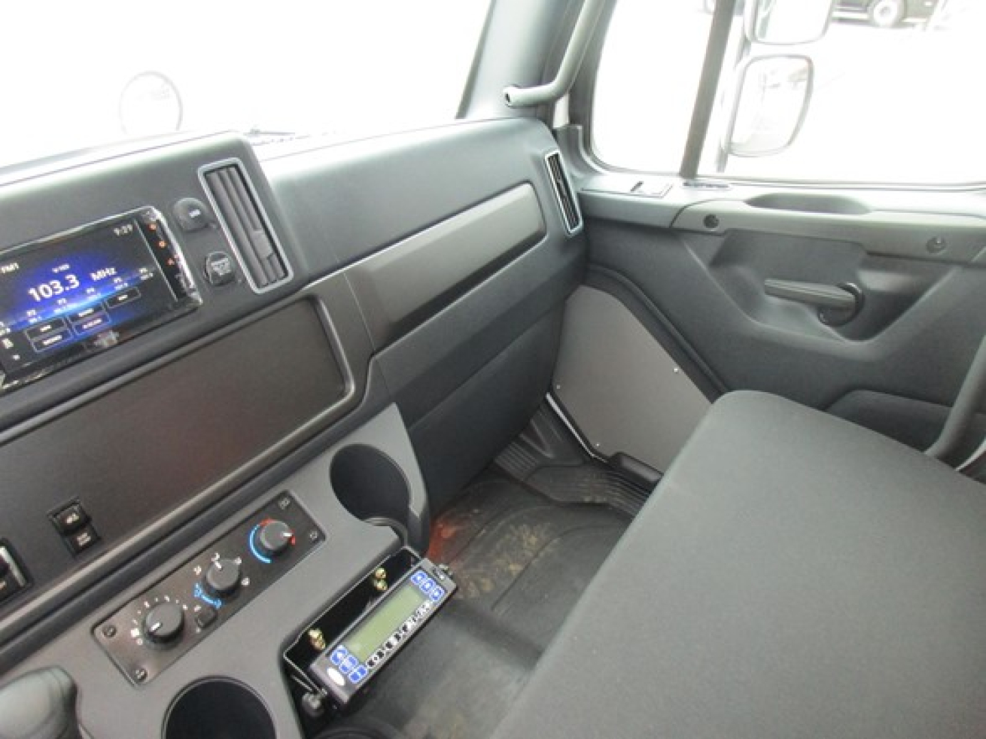 Freightliner M2 106 Plus cab interior showing steering area, dashboard with radio display, climate controls, and storage compartments.
