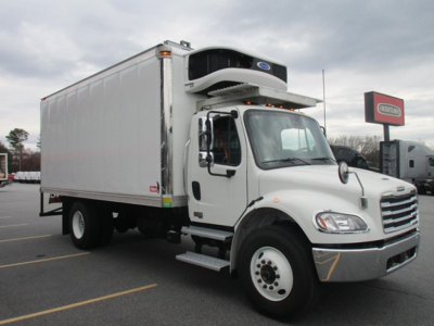 Freightliner M2 106Plus refrigerated truck in a dealership lot, side view with cargo box and APU unit on top, white cab and chassis.