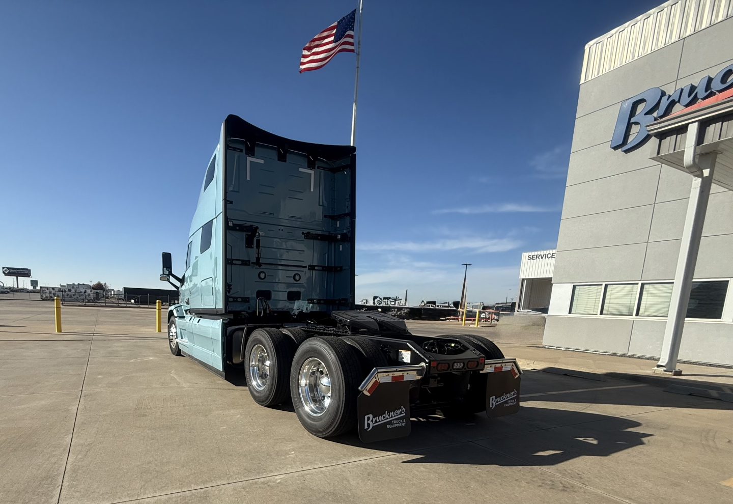 Truck tractor rear three-quarter view showing fifth-wheel and dual rear axles, light blue cab, tall sleeper, chrome wheels, and Bruns truck dealer building in background.