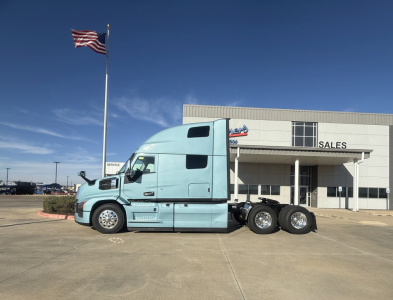 Fleet truck: a light blue 2026 Mack PR64T76H conventional sleeper tractor parked in front of a sales building with an American flag nearby.