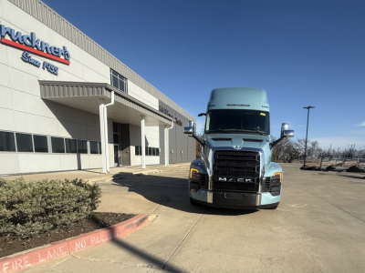 Front view of a blue 2026 Mack PR64T76H truck in a dealership lot with a building and sign in the background.