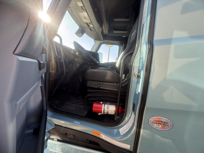 Truck cab interior of a 2026 Mack PR64T76H with front door open showing steering wheel, dashboard, and a fire extinguisher mounted near driver seat.