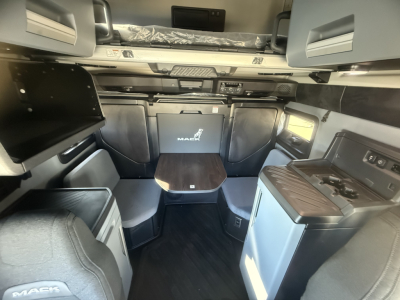 Truck cab interior showing sleeper with bunk above, compact galley area with countertop and sink, table in center, and seating around; interior branded with Mack logo on cabinet panels.