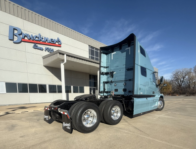 Mack PR64T76H conventional sleeper truck in blue parked outside dealer, rear view shows tandem axles and fifth wheel, landing gear, and Bruchner's dealership building in background.