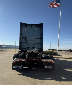 Rear view of a 2026 Mack PR64T76H conventional sleeper truck chassis with extended cab and dual rear wheels; flagpole with American flag in background; mud flaps say Bruckner Equipment.