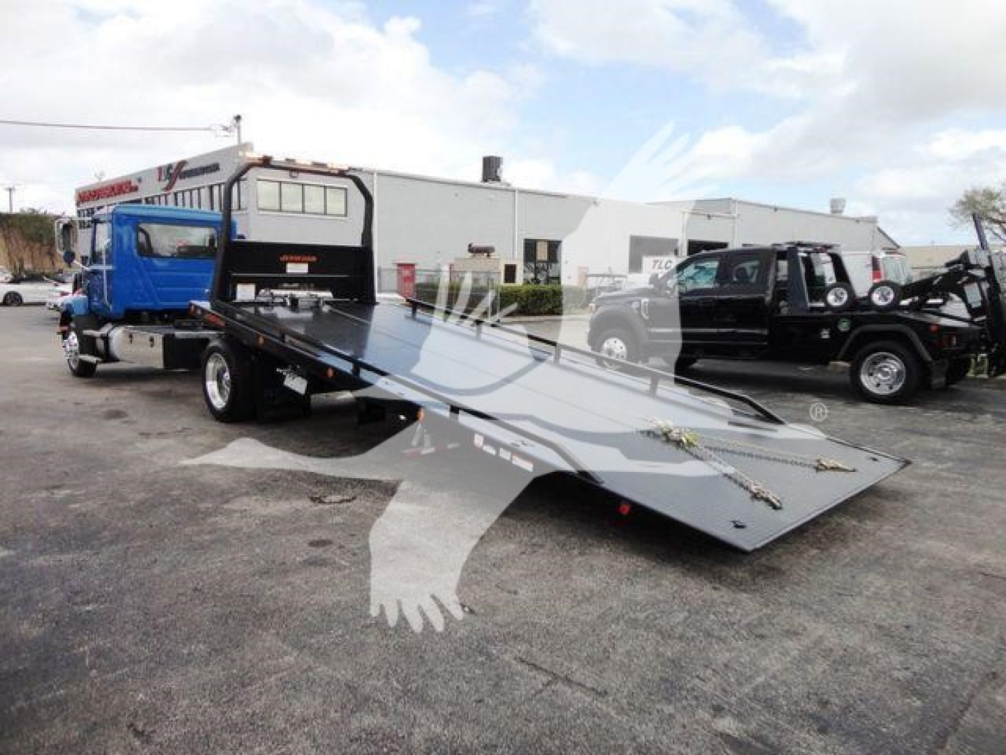 Truck with a flatbed rollback tow truck in a parking lot, Mack MD6 front view with extended ramp and heavy-duty tilt bed, dealership backdrop.