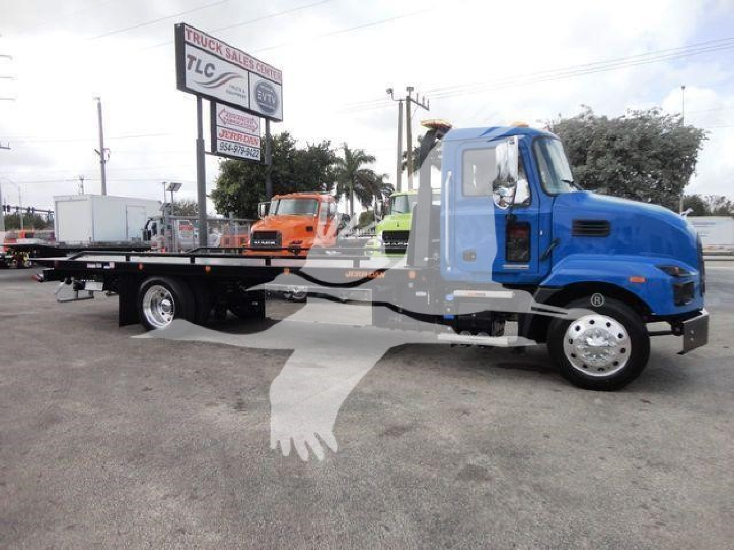 Blue Mack MD6 truck with a long flatbed rollback body in a dealership lot, cab interior visible through open window, other trucks in background.