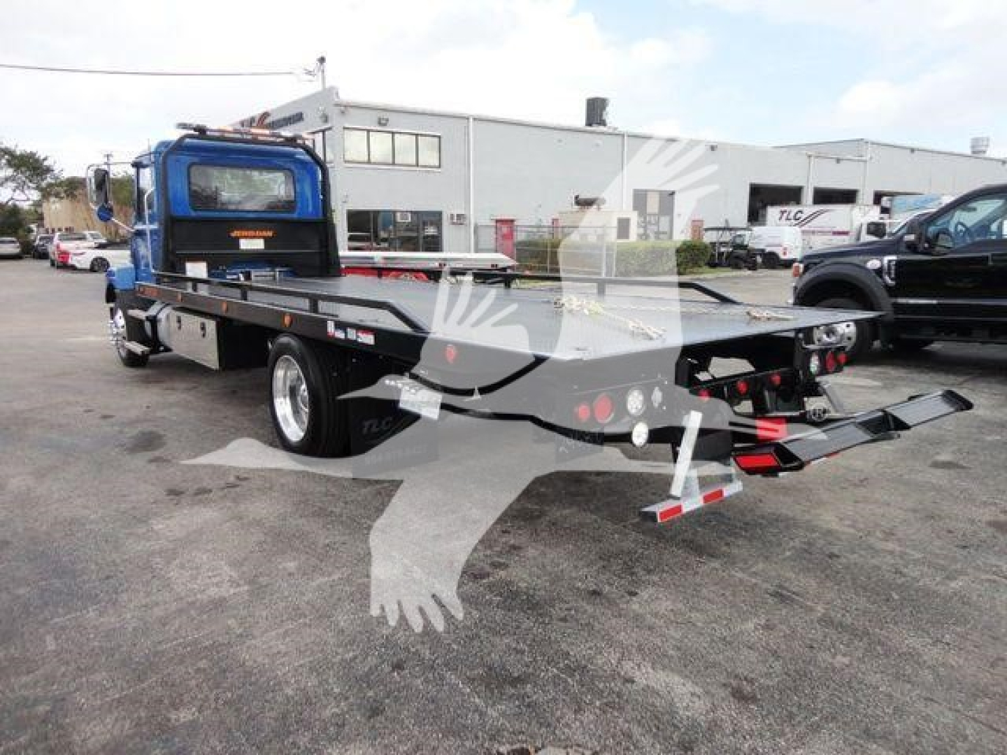 Flatbed rollback tow truck with blue cab and black flatbed showing hydraulic ramps and rear lights.