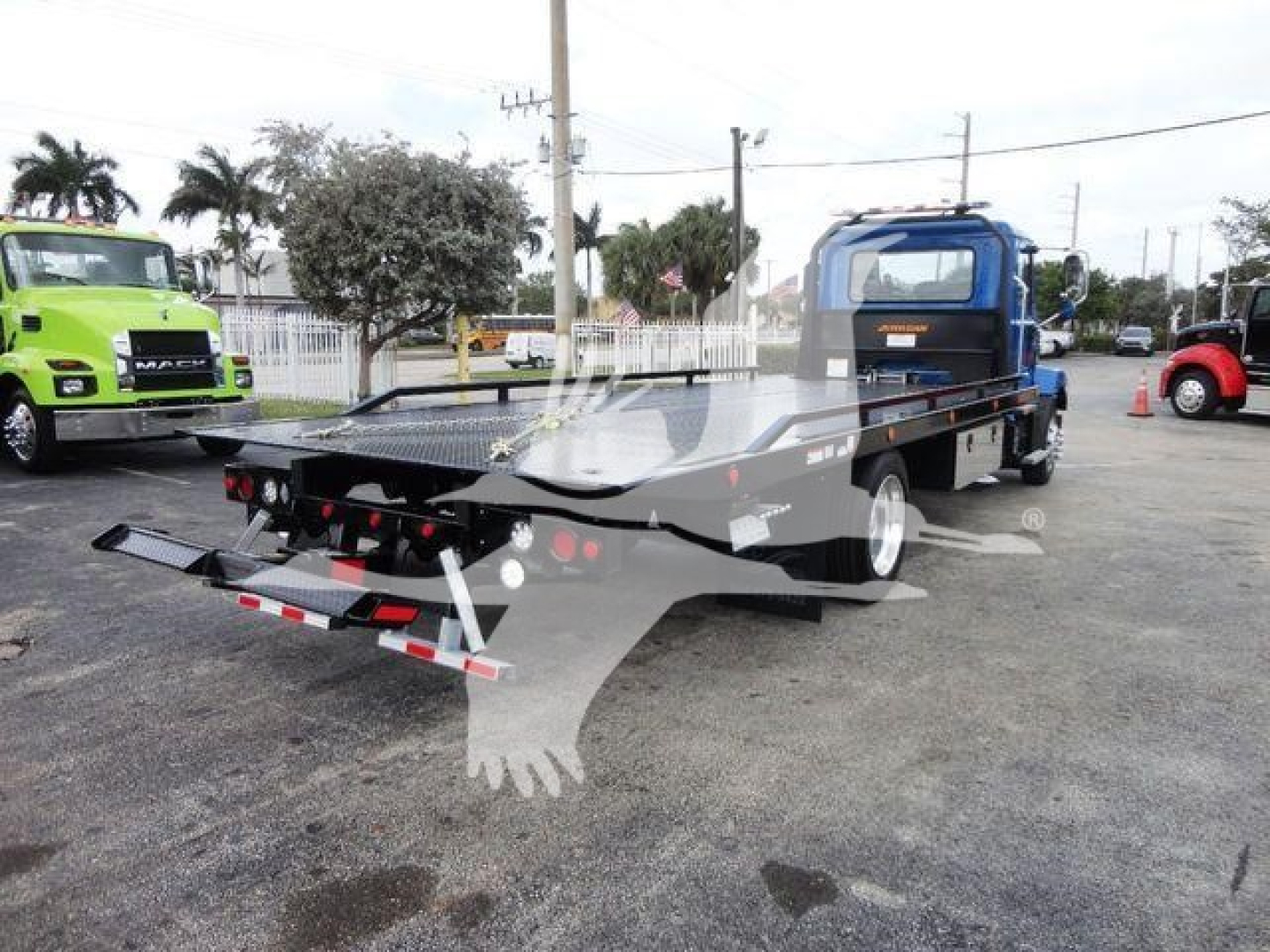 Flatbed tow truck with blue cab and extended platform, rear ramp and wheel chocks visible; Mack MD6 style truck in a dealership lot.