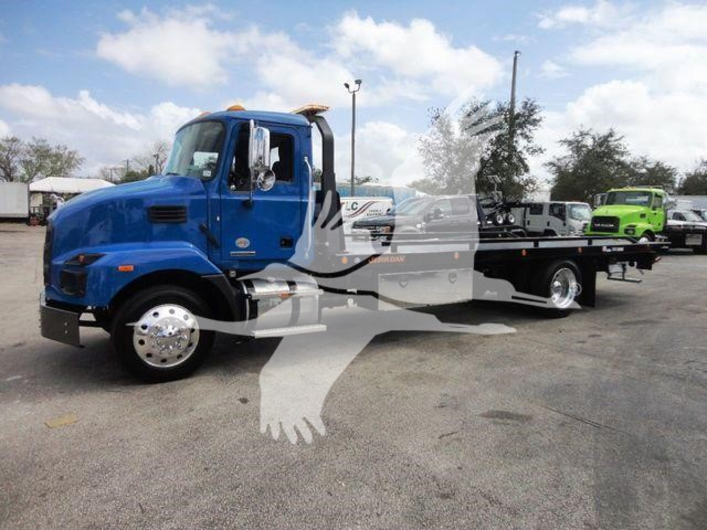 Blue Mack MD6 tow truck in a lot with flatbed and another green truck in the background.