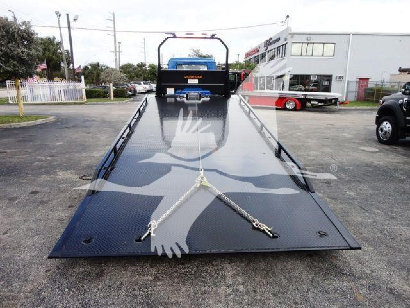 Flatbed tow truck deck on a new Mack MD6 rollback trailer with a blue winch and chains secured. Stock number J04369 visible in background; dealership lot.