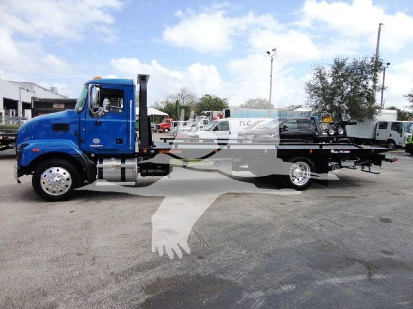 Blue Mack MD6 rollback tow truck in a lot showing cab and long flatbed with wheel lift equipment and chrome exhaust stack.