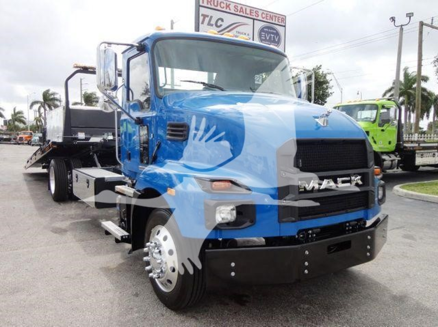 Blue Mack MD6 rollback truck in a dealership lot showing front view, grille with MACK emblem, large headlights, black bumper, side mirrors, and flatbed behind.