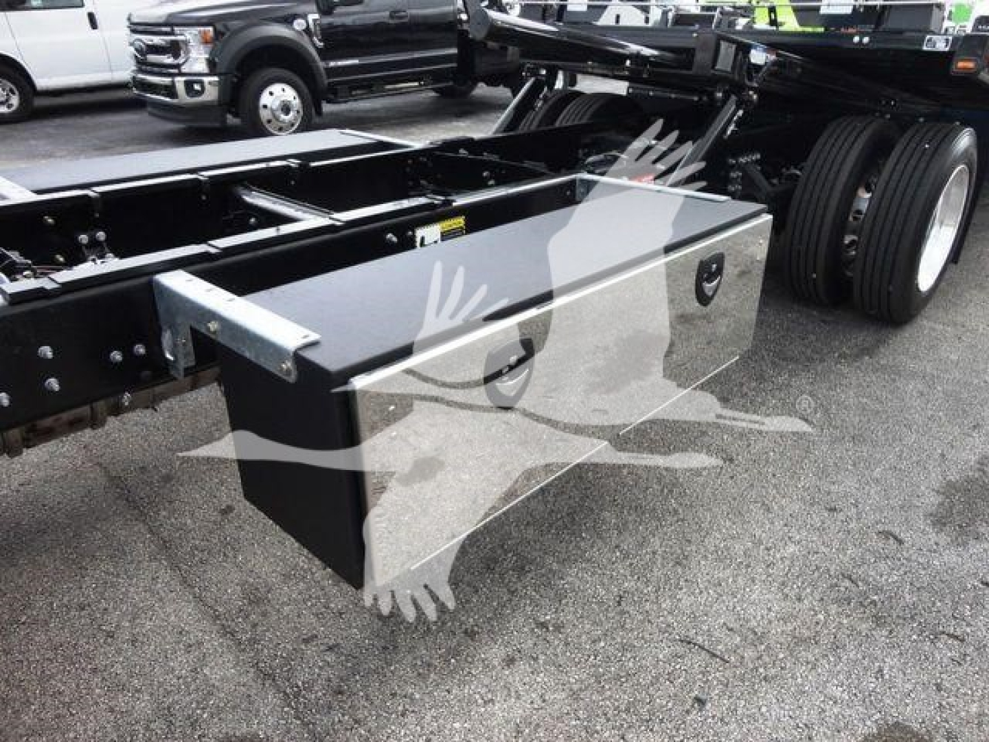 Truck chassis with a large metal tool/storage toolbox mounted on the frame, showing a black rectangular box with two lidded compartments and a step area nearby.