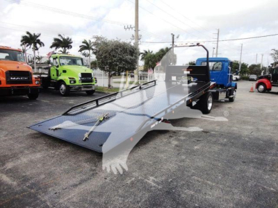 Flatbed rollback truck with lowered ramp in foreground; Mack MD6 dealer lot with colorful trucks in background.