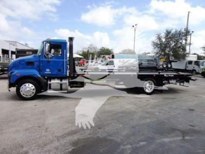 Blue Mack MD6 rollback tow truck in a lot showing cab and long flatbed with wheel lift equipment and chrome exhaust stack.