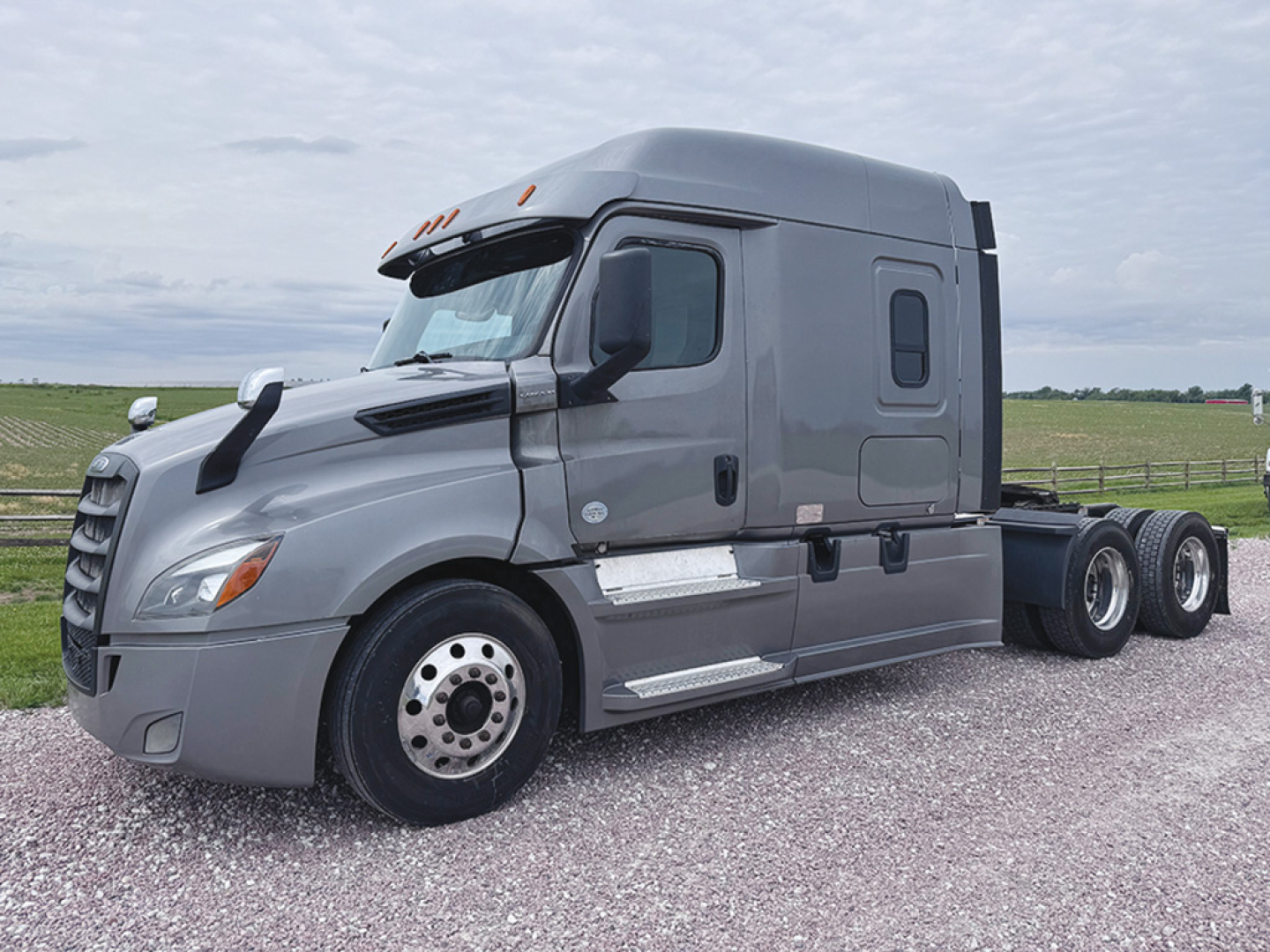 Truck cab of a 2023 Freightliner Cascadia126 in a gray color, side view showing sleeper cab with window, mirrors, and dual rear axles.
