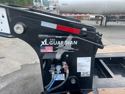 Truck trailer hitch component with XL Guardian logo, American flag, and hydraulic hoses. Visible text includes THE XL GUARDIAN, DO NOT SIDE LOAD, NOTICE, blue hoses connected to fittings, black metal frame.