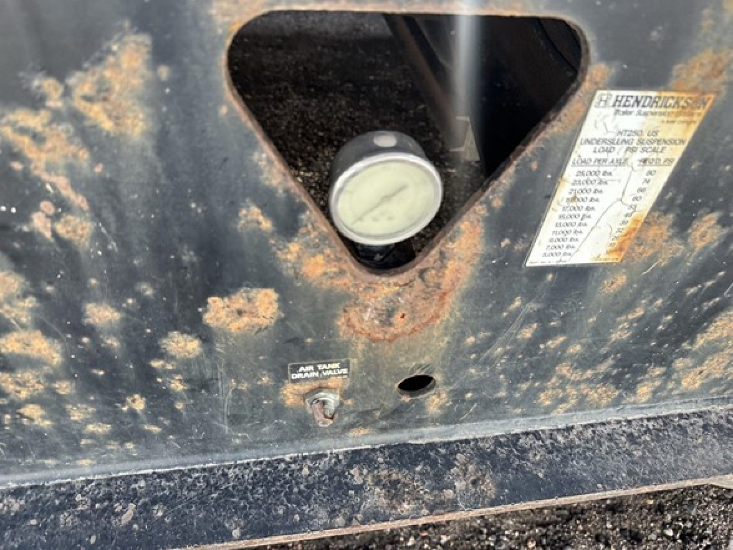 Trail King 40 ton hydraulic tail trailer label and gauge visible on black metal surface with rust specks and a warning label. Round gauge window, stickers with specs, and a small bolt.