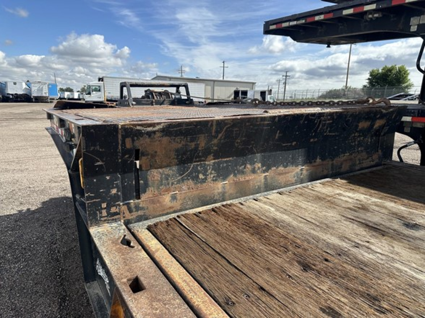 Flatbed trailer deck with wooden planks and rusted steel side rails, tail section visible, parked in lot with other trailers.