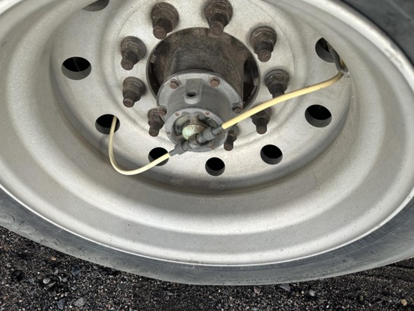 Wheel hub and lug nuts of a trailer axle with a yellow wire/strap near the hub, showing a steel rim and multi-hole pattern.
