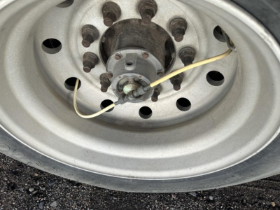 Wheel hub and lug nuts of a trailer axle with a yellow wire/strap near the hub, showing a steel rim and multi-hole pattern.