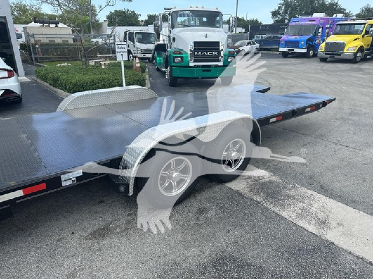 Trailer bed and rear wheel area showing flatbed tilt with diamond-plate surface; visible marquees and other trucks in background.