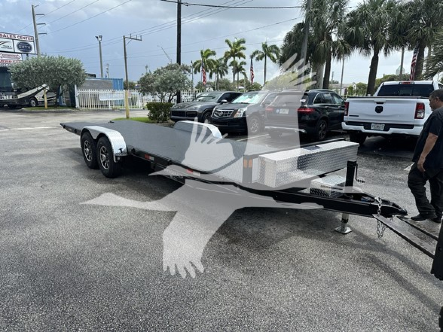 Trailer in a parking lot showing a flatbed utility trailer with dual rear axles and a diamond plate toolbox near the hitch.