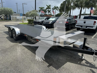 Trailer with low-profile flatbed and dual rear axles, aluminum diamond plate walkway, and front toolbox on a dealership lot.