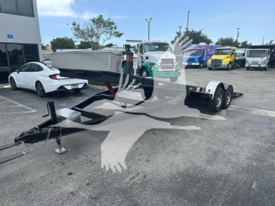 Trailer hitch and dual-axle flatbed trailer in a dealership lot with other trucks, showing a V-shape frame and a long bed.