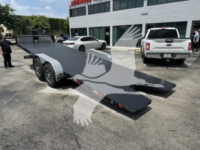 Trailer showing flatbed car hauler with dual axles and rear ramp sections, parked in lot in front of a white building.