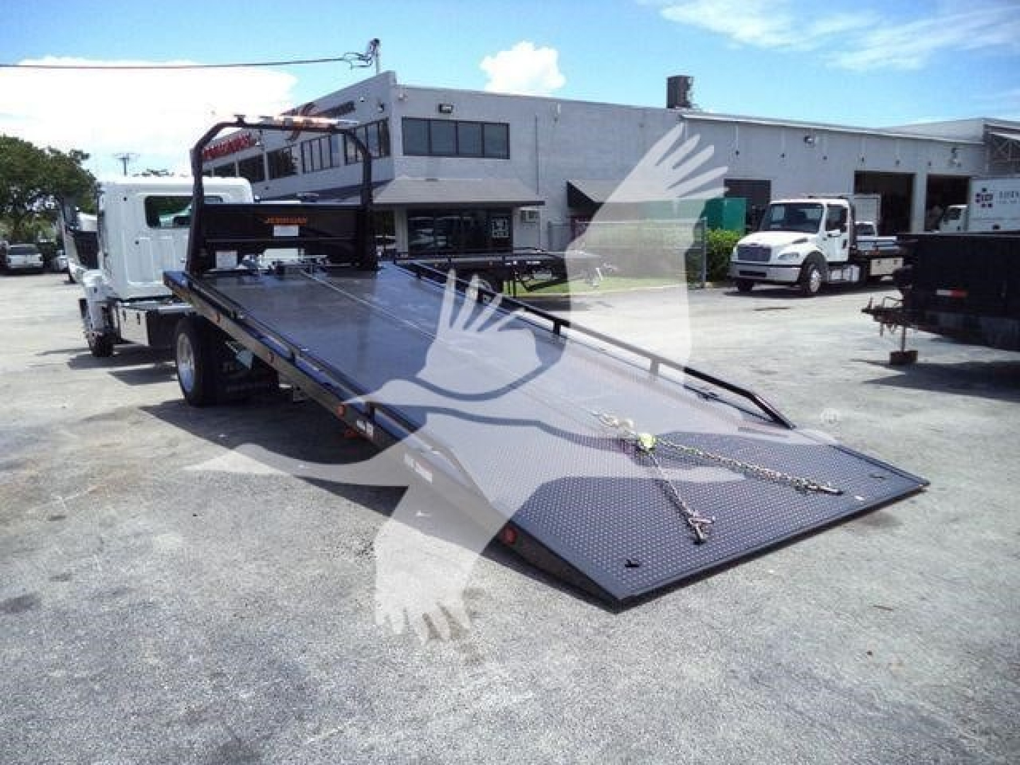 2026 Hino L6 rollback carry flatbed tow truck with ramp lowered and hydraulic deck, showroom area in background with other trucks.