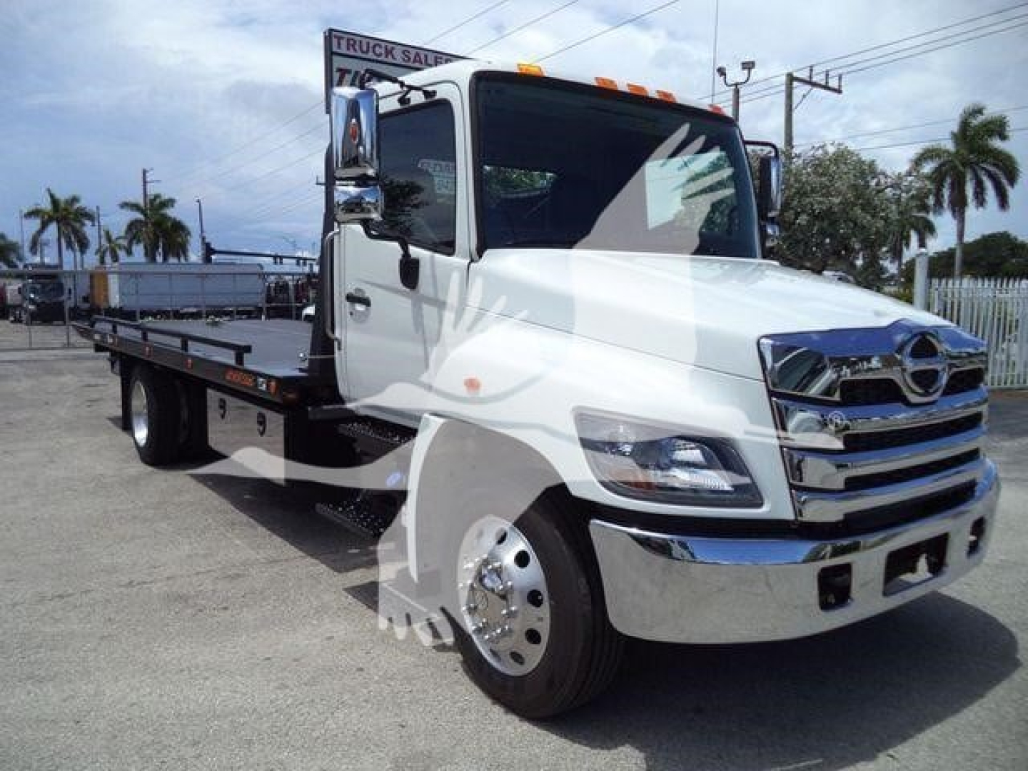 Flatbed tow truck with white cab and long black bed, chrome grille, Hino badge visible, side mirrors, and orange clearance lights on cab roof.