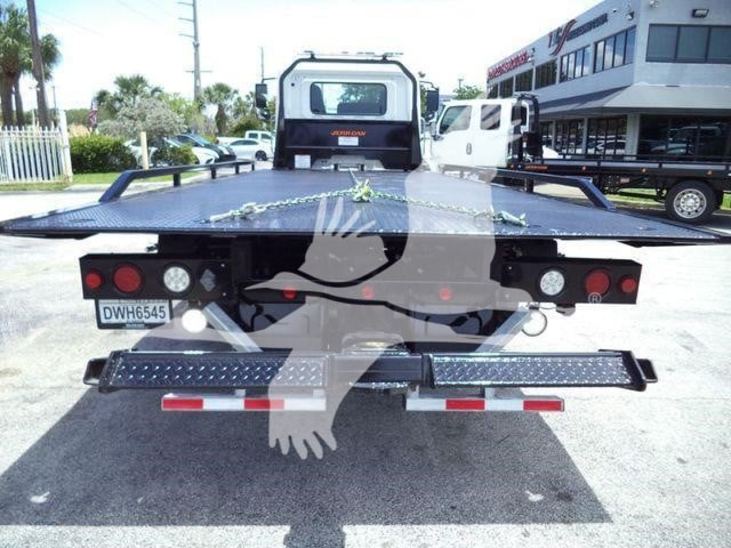 Rear view of a flatbed tow truck with taillights, license plate DWH6545, and metal steps; black flatbed with chain, and a white cab in background.