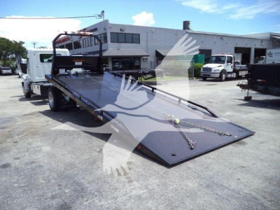 2026 Hino L6 rollback carry flatbed tow truck with ramp lowered and hydraulic deck, showroom area in background with other trucks.