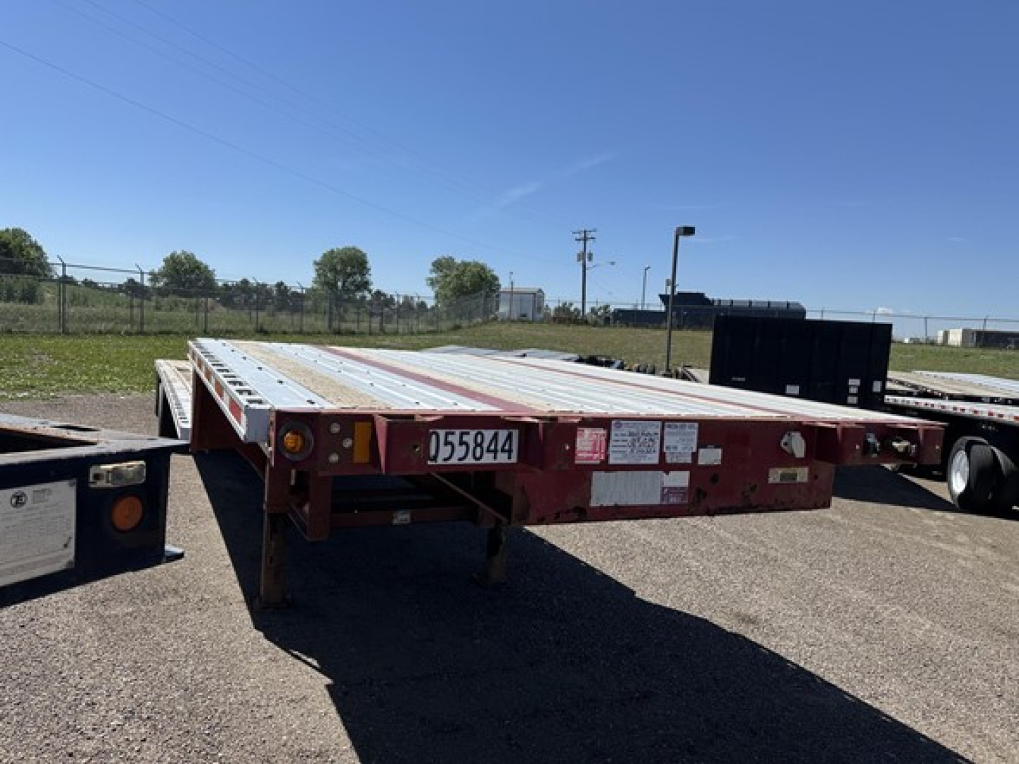 Flatbed trailer with drop deck in a parking lot showing side rails and rear frame; visible stickers and serial plate.